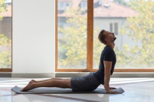 A person practicing yoga poses on a mat beside large windows, surrounded by natural light and greenery outside.