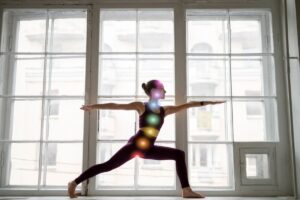 A person practices yoga in a warrior pose, stretching their arms, with large windows and a blurred urban backdrop.