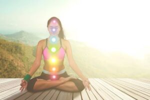 A person meditates in a yoga pose on a wooden deck, with colorful chakra symbols glowing along their spine against a serene landscape.