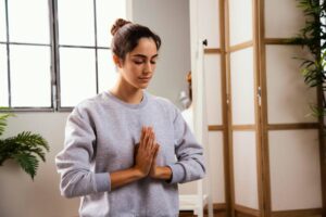 A woman in a grey sweatshirt performs a yoga pose with her hands in a prayer position, surrounded by indoor plants and natural light.