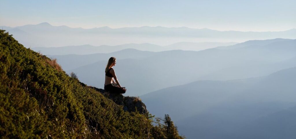 A woman sits in a meditative pose on a cliff, overlooking misty mountains under a clear sky, embodying tranquility and connection with nature.