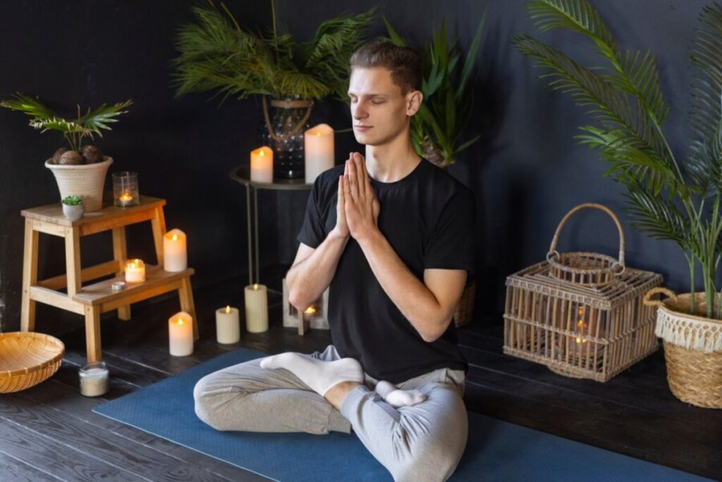 A person sits in a meditative pose on a yoga mat, surrounded by plants and softly glowing candles in a calming interior space.