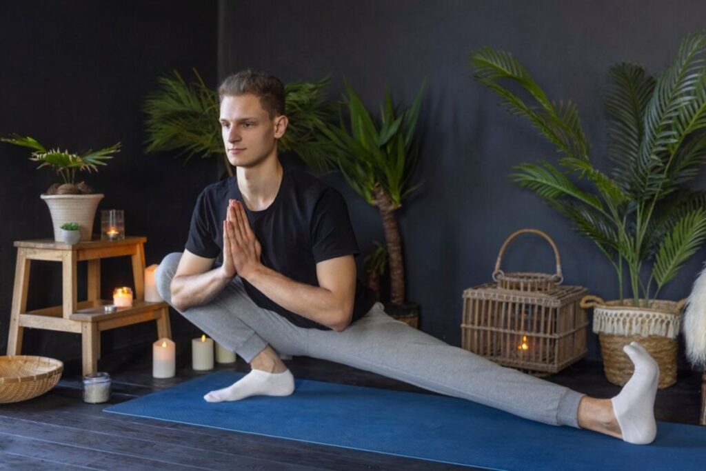 A person practices yoga on a blue mat surrounded by plants, candles, and wooden decor in a serene indoor space.