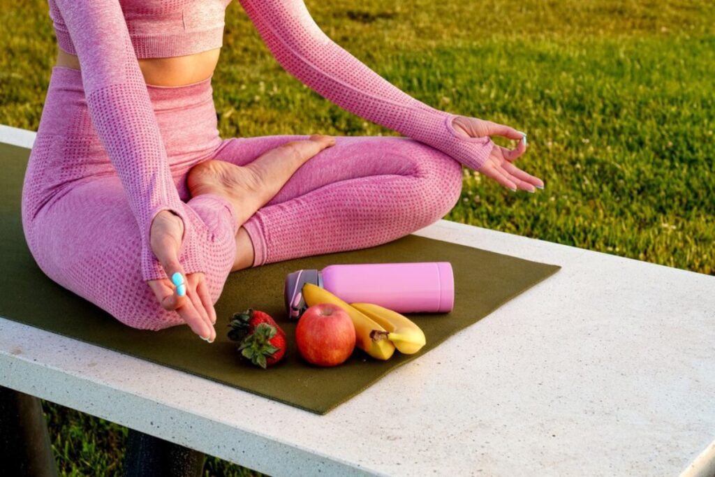 A woman in pink workout attire meditates on a mat, surrounded by fruit and a water bottle, against a green grassy background.