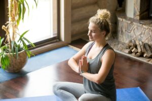 A person sits cross-legged on a yoga mat, practicing meditation with hands in a prayer position, surrounded by natural light and greenery.