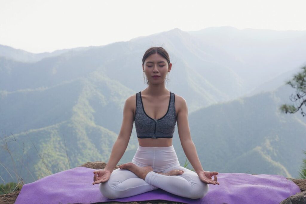 A person meditating in a cross-legged position on a purple mat, surrounded by serene mountain scenery.