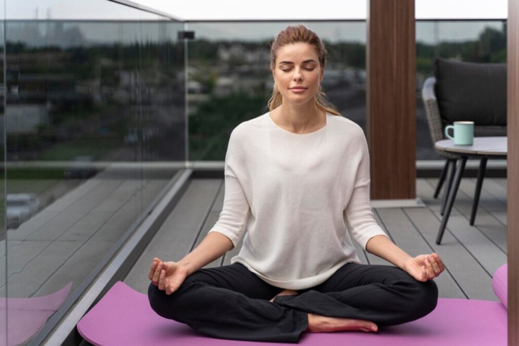 A person is meditating on a purple yoga mat on a balcony, surrounded by a modern setup and urban scenery in the background.