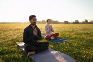 Two individuals practice yoga in a sunlit field, seated on mats, surrounded by blossoming flowers and a serene landscape.