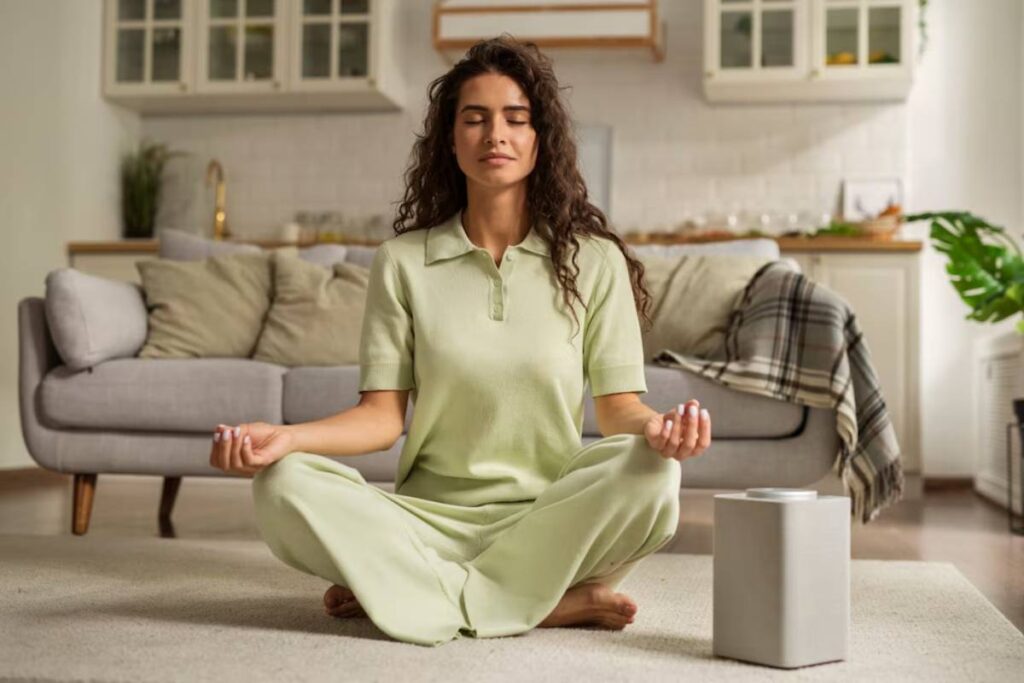A woman in a light green outfit sits cross-legged in a serene living room, practicing meditation beside a modern speaker.